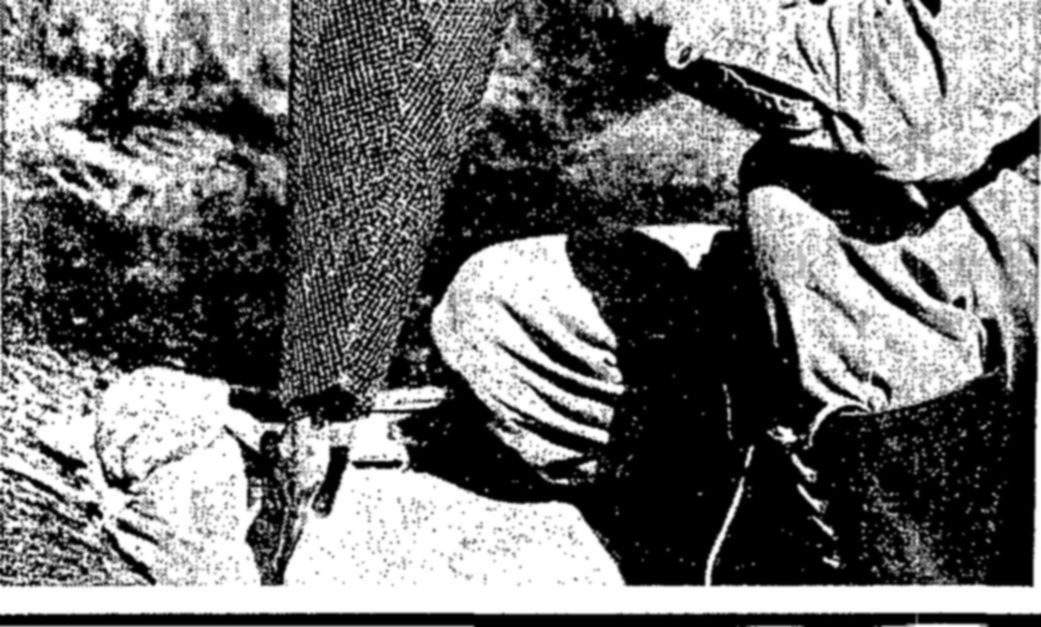 Coarse, tightly cropped photograph showing the legs and boots of a crouching person beside a long dark object, possibly a net or pole, and a cloth bundle on the ground.