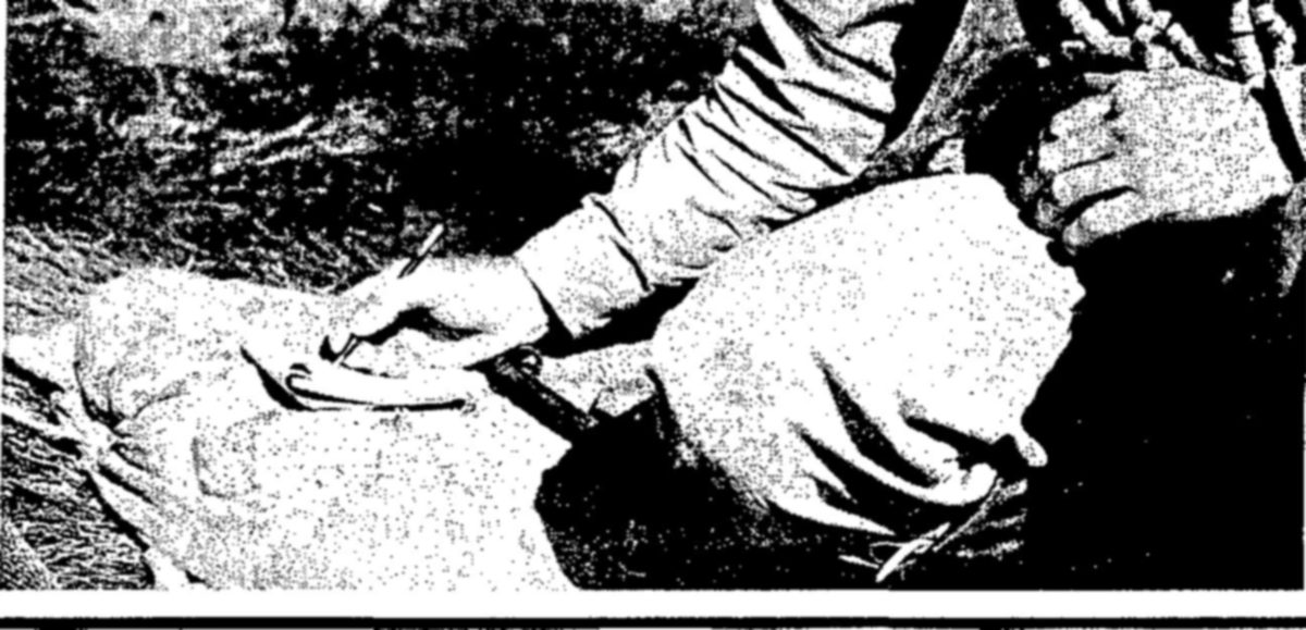 Tightly cropped photograph of a crouching person in work clothes writing with a pencil on a pad or cloth on the ground, with the other hand held up at the right.