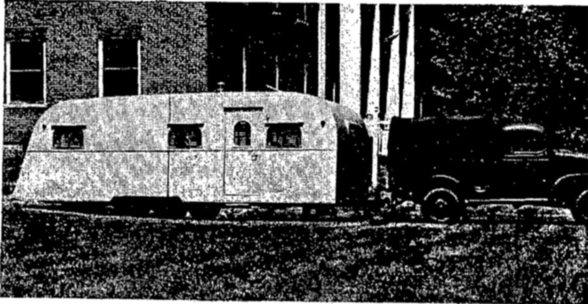 A rounded-roof house trailer with several small windows and a door, hitched to a dark truck, parked on grass in front of a brick building.