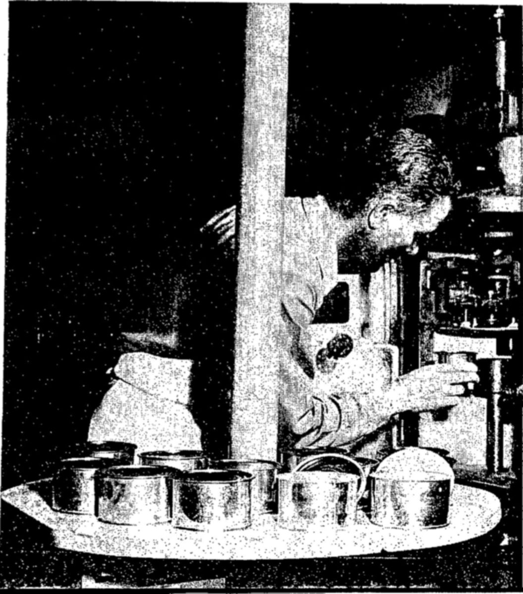 A man leans toward a small machine, working a lever with one hand, while a round table in front of him holds a ring of open metal cans.