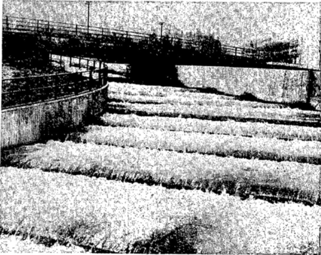 Water flows down a stepped concrete channel in a series of white-topped ridges between high walls, with a railed walkway or bridge crossing above.