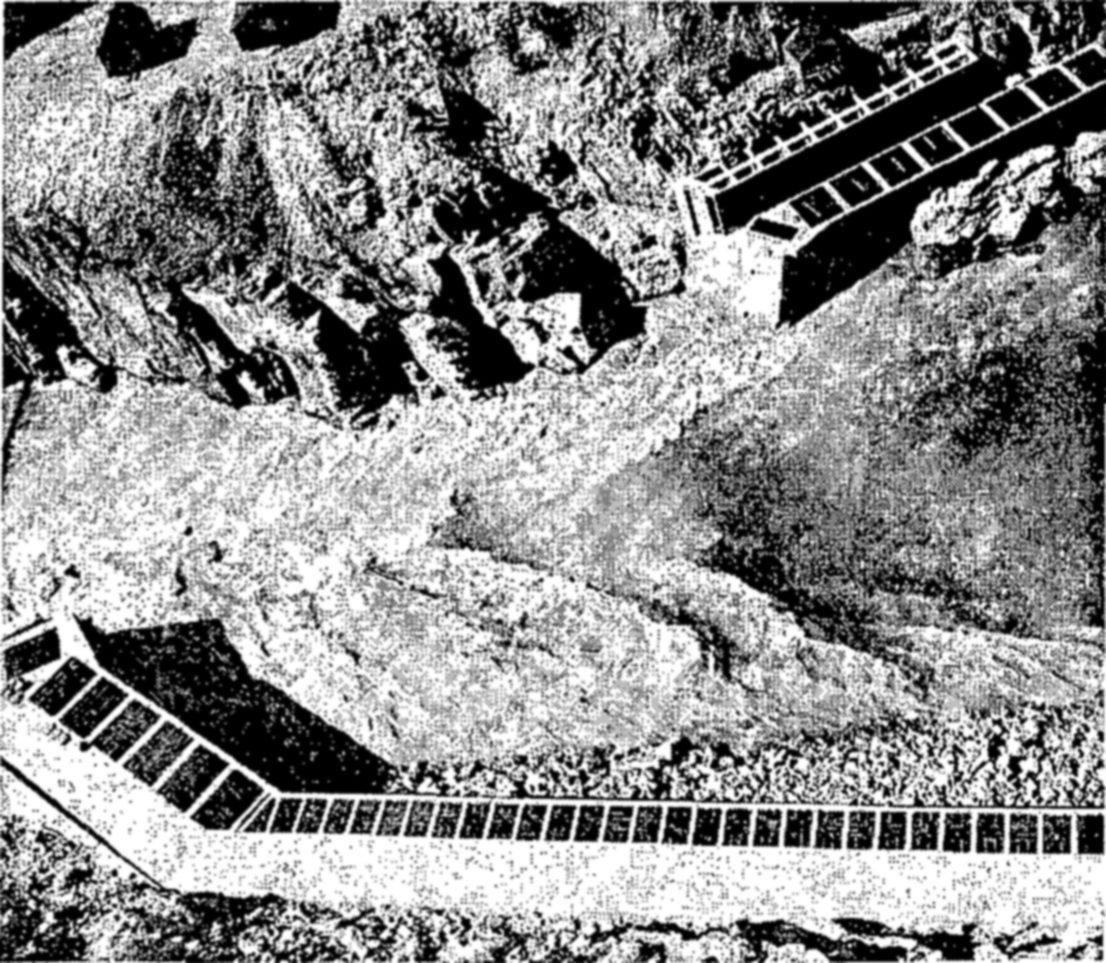 Fishways at Hell’s Gate in the Fraser River canyon. — Seen from above, long concrete channels divided into square compartments run along both sides of a rocky canyon with rushing water between them.