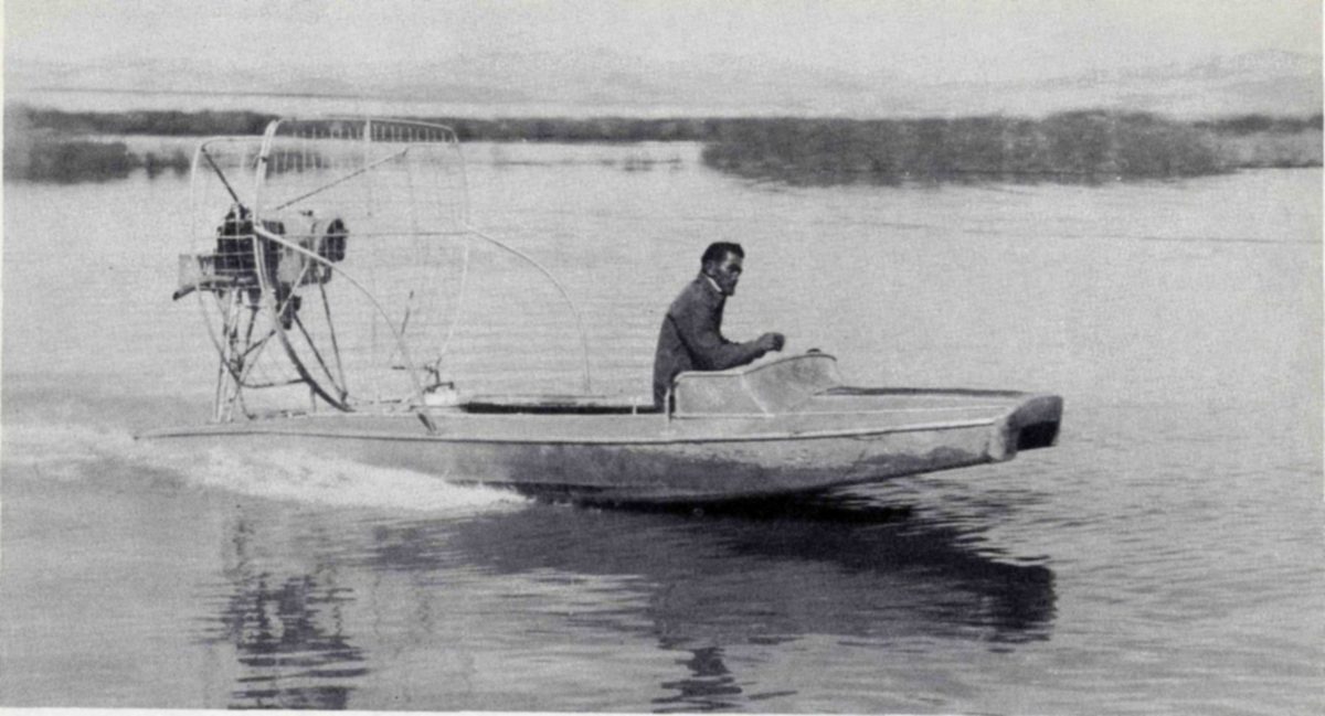 The air boat, with its flat-bottomed aluminum body and airplane propeller, was developed at Bear River to allow easy and rapid travel on the very shallow waters over the mud flats. Now this type of boat is used in many parts of the country for travel in marshes and other still, shallow waters. — Photograph of a man steering a low flat boat across open water, with an engine and a large propeller inside a wire guard mounted at the rear and a reedy shore in the distance.