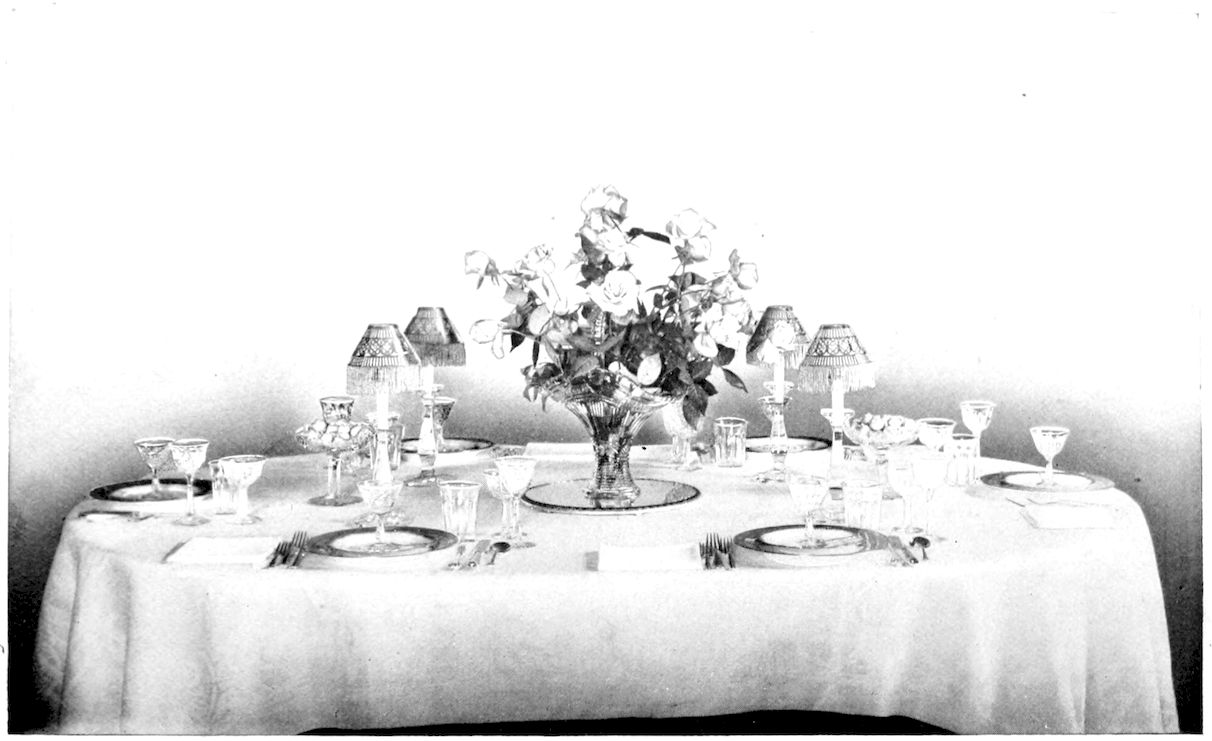 Table laid for Formal Dinner. — Frontispiece. — A table under a white damask cloth, with a cut-glass vase of roses on a round mirror at the centre, four candles under fringed shades, and plates, silver and wine glasses at each cover.