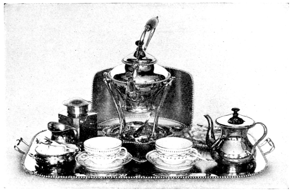Five o’Clock Tea Service. — A beaded silver tray holding a tea-kettle hung over its spirit lamp, a tea caddy, cream jug, sugar bowl, a teapot, and two china cups on saucers.