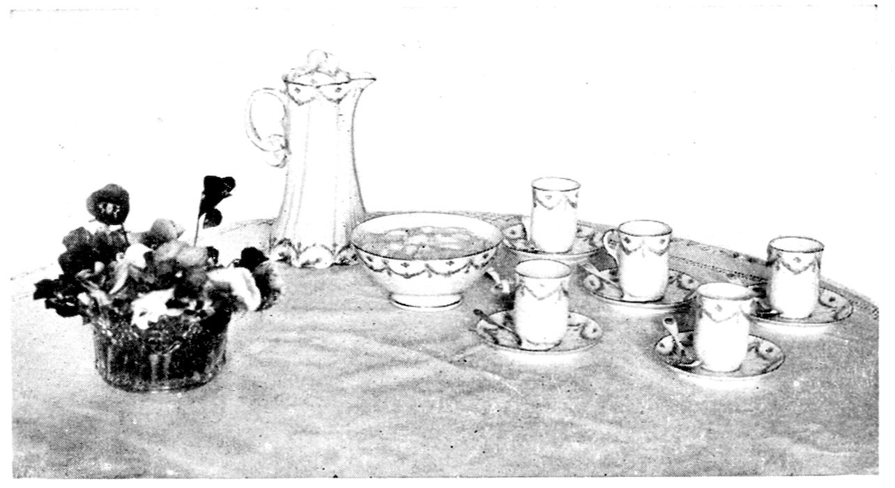 Chocolate Service. — On a cloth-covered table: a tall china chocolate pot, a bowl of whipped cream in the same pattern, five tall chocolate cups on saucers with spoons, and a low glass bowl of pansies.