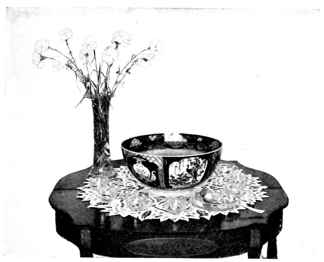 Punch Service. — A large decorated punch bowl stands on a lace doily on a small table, with punch glasses and a ladle around it and a tall glass vase of carnations beside it.