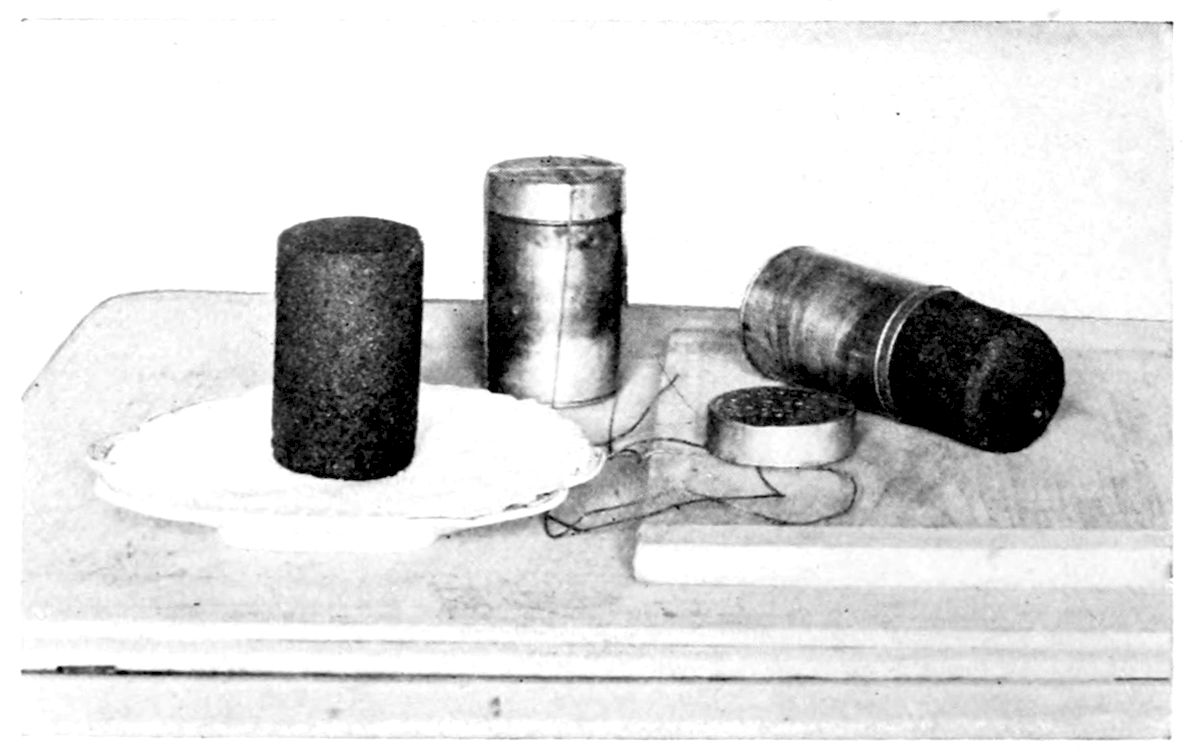 Boston Brown Bread. — A steamed loaf of Boston Brown Bread turned out on a plate, beside two baking-powder boxes used as moulds, one closed and one on its side showing the bread, a lid, and string.