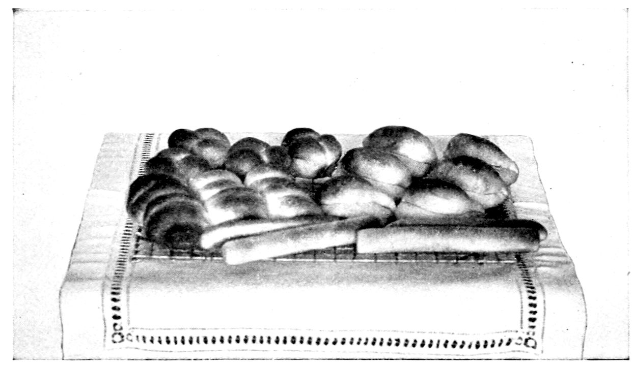 Parker House Rolls; Salad Rolls; Clover Leaf Biscuit; Sticks. — Rows of browned rolls on a wire rack set on a hemstitched cloth: folded and creased rolls, three-lobed clover leaf biscuits at the back, and long smooth sticks laid across the front.