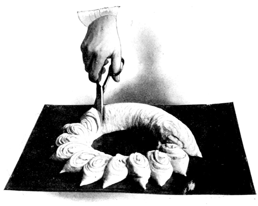 Swedish Tea Ring II before baking. — A hand with scissors snips a long roll of dough, joined into a ring on a baking sheet; the slices already cut are turned outward, spiral side up, round half of the ring.