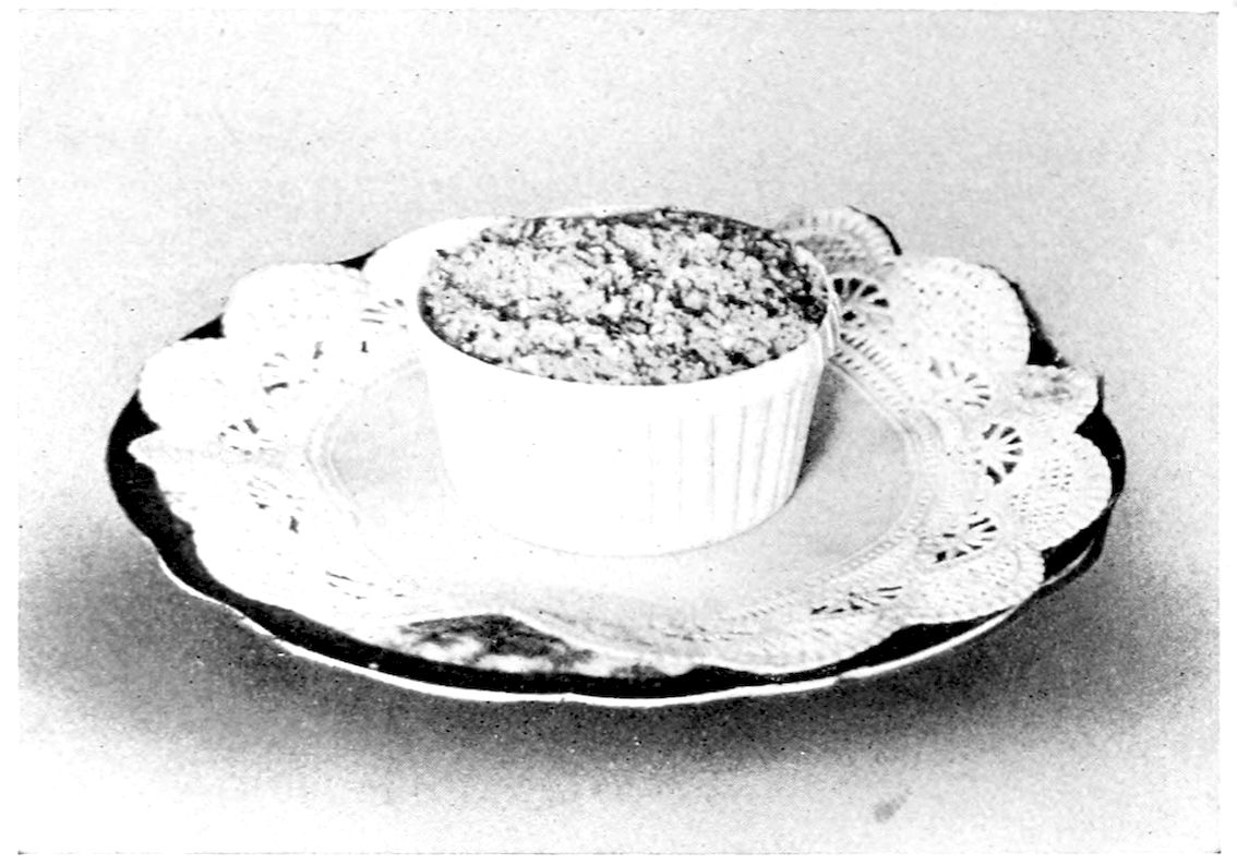 Shirred Egg. — A single fluted white shirrer on a paper doily, its egg hidden under a browned layer of crumbs.