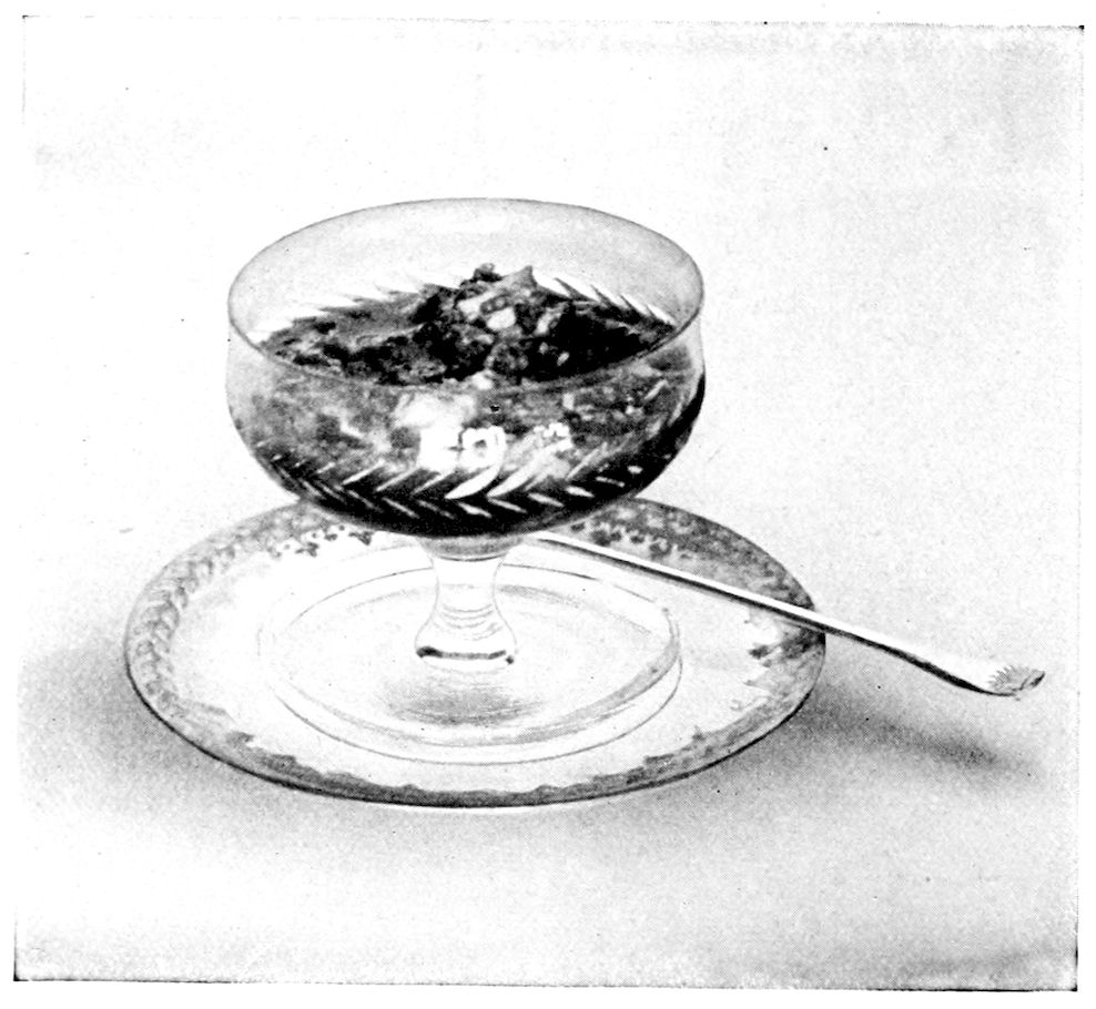Lobster Cocktail. — The lobster meat is heaped in a stemmed cocktail glass cut with a band of leaves, set on a glass plate with a long-handled spoon beside it.