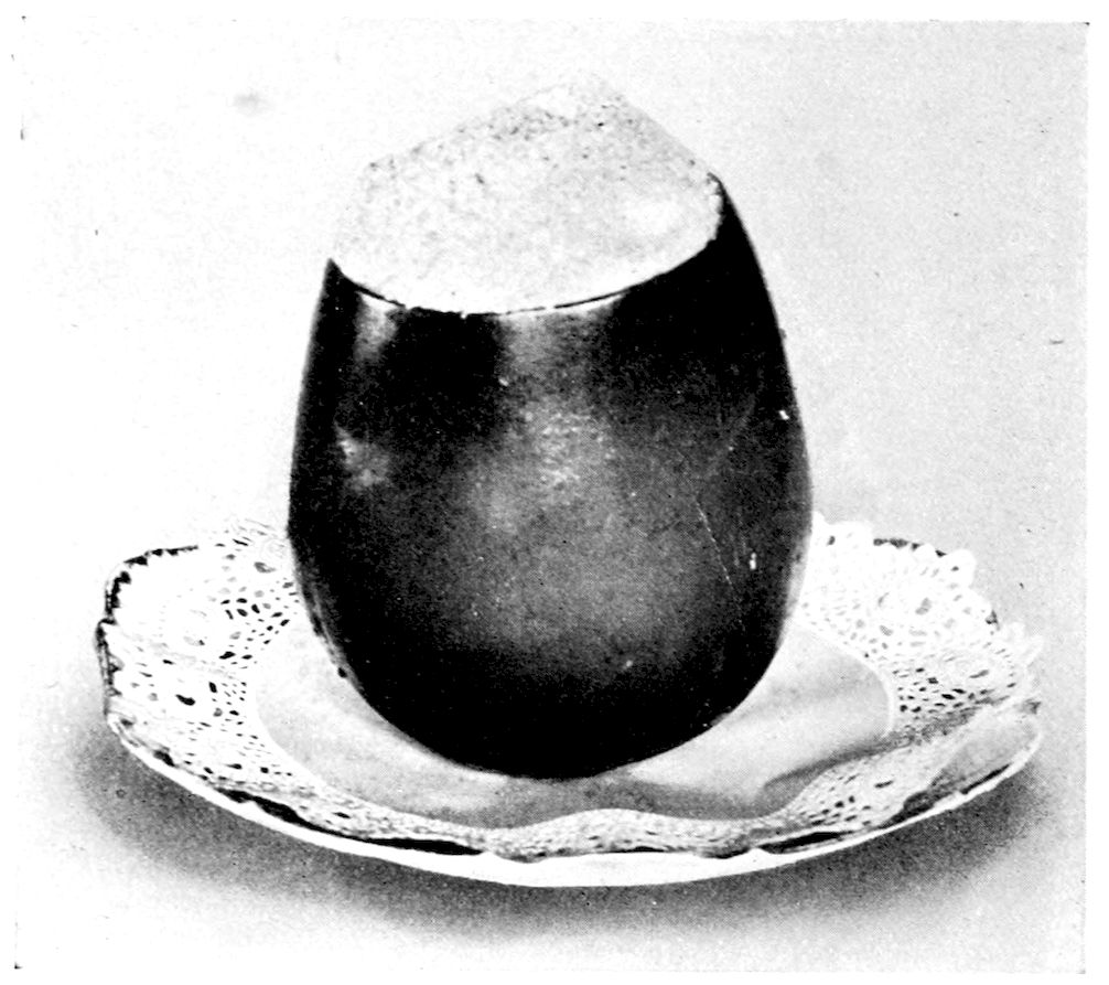 Stuffed Egg Plant. — The whole eggplant stands upright on a lace doily on a plate, its dark glossy skin intact and its cut top heaped with a browned crust of buttered crumbs.