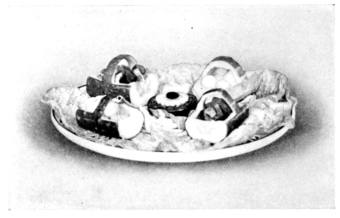 Cucumber Baskets. — Cucumber halves cut into baskets with arched handles sit on lettuce leaves on an oval platter, some holding pale cubes and some darker pieces, with a round slice at the centre.