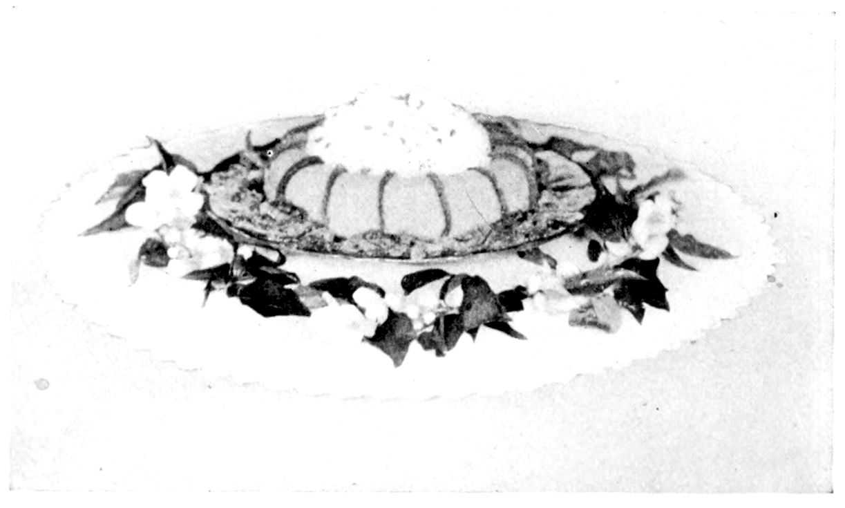 Orange Trifle garnished with Whipped Cream, Candied Orange Peel, and Blossoms. — A fluted ring mould with thin strips of candied peel along its ridges and whipped cream heaped in the centre, ringed by a wreath of leaves and orange blossoms on a doily.