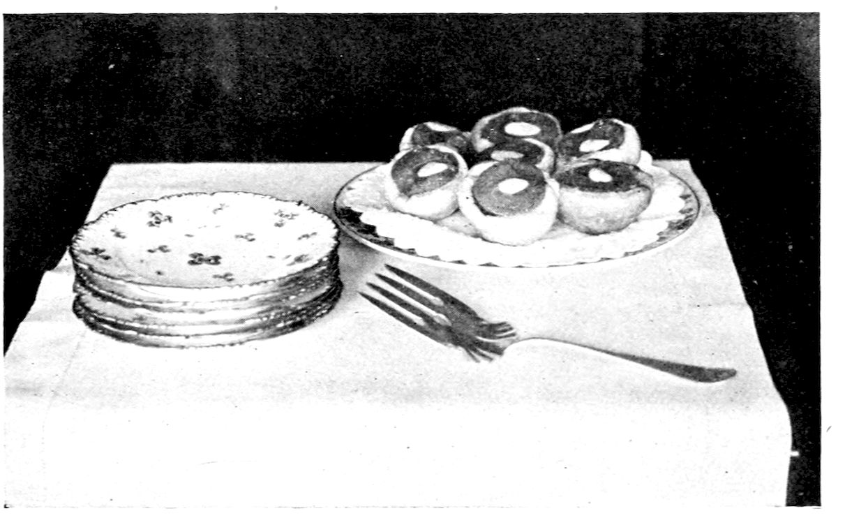 Calvé Tarts. — Seven baked tart cases, each holding half a peach with a blanched almond at its centre, on a doily-lined plate, with a stack of small plates and a fork beside them.