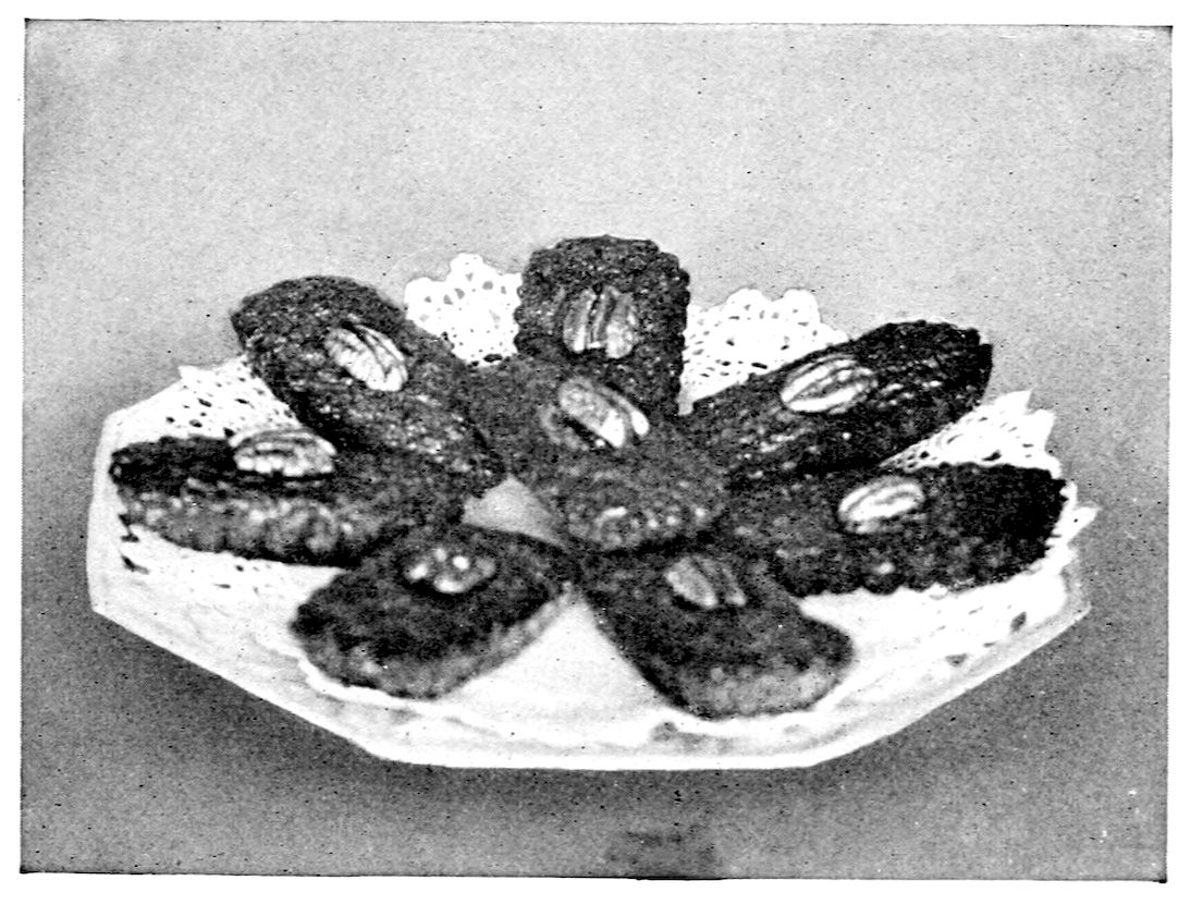 Marguerites I. — Eight small oblong cakes, each topped with a pecan half, are laid in a star with their ends meeting at the centre, on a lace doily on an eight-sided plate.