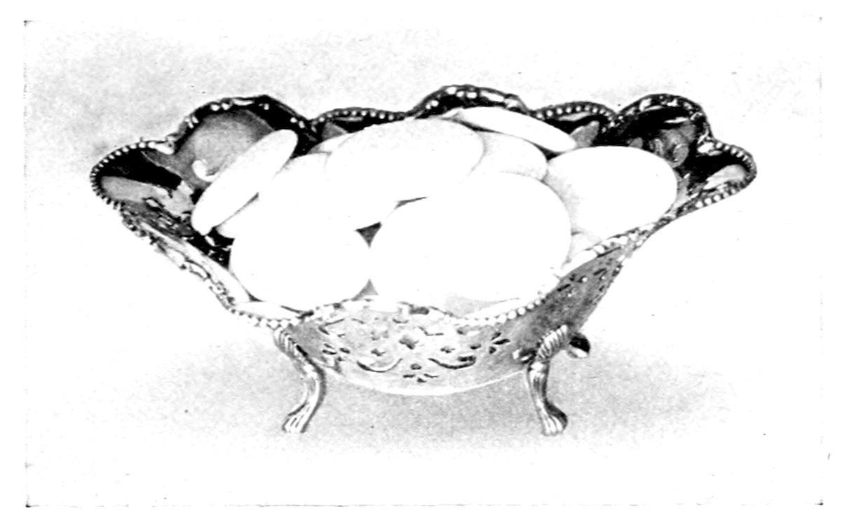 Cream Mints. — Flat round cream mints, pale and smooth, fill a small silver bonbon dish with a beaded, scalloped rim, a pierced band and three curved feet.