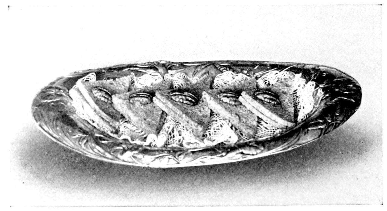 Noisette Sandwiches. — Triangles of dark nut bread, each with a walnut half pressed on top and a thin line of filling showing at the edge, stand in a row on lace in an oval embossed silver dish.
