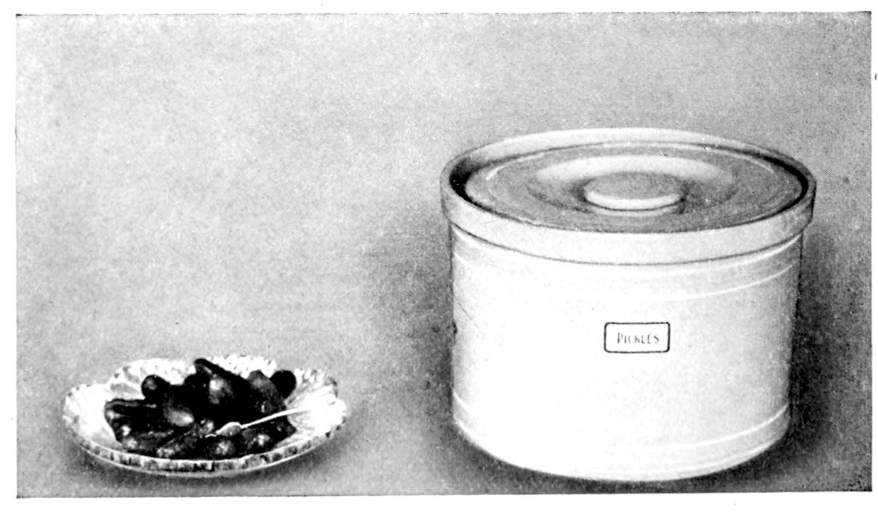 Pickles ready for serving. Crock for keeping Pickles. — A small cut-glass dish of dark little pickles with a fork beside it, and a large covered stoneware crock labelled Pickles.