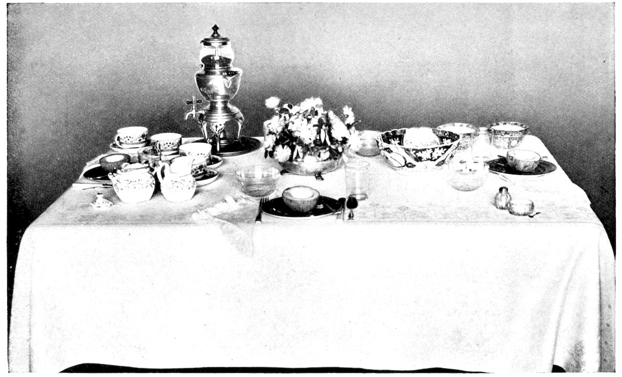 Table laid for Breakfast. — A breakfast table with a coffee urn at one end among cups, saucers, a cream jug and sugar bowl, a low bowl of flowers in the centre, and a single place laid with a halved fruit on a plate.