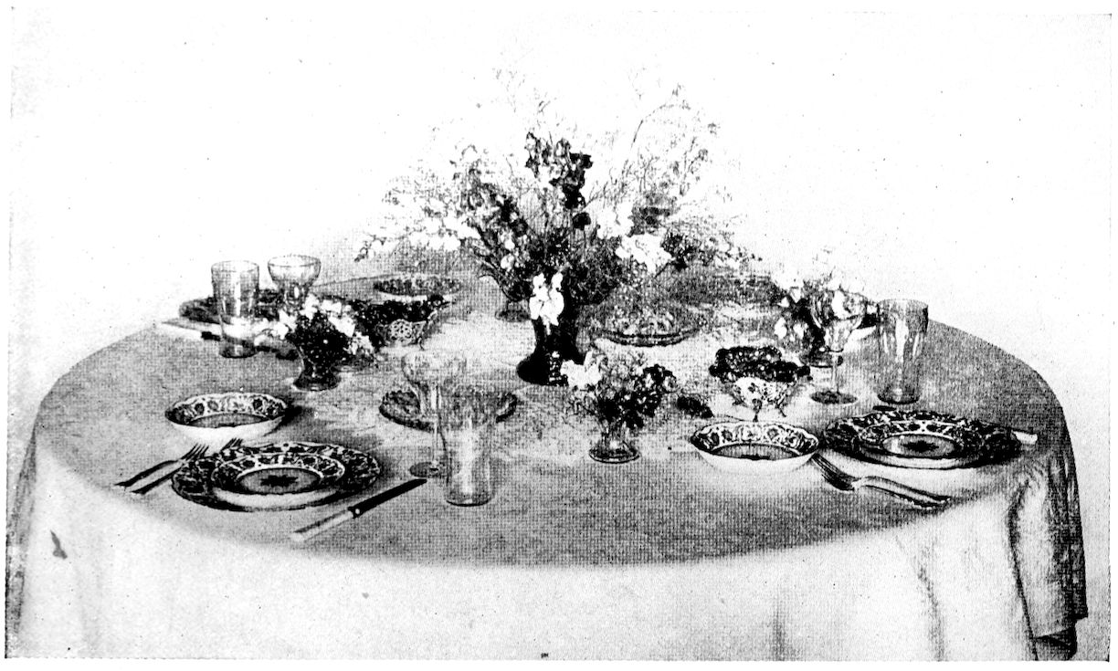Luncheon table laid for Fish Course, Soup Course presumably having been removed. — A round table laid with patterned plates and a small bowl at each place, tumblers and small glass dishes, small posies in low vases between the places and a tall spray of flowers in the centre.