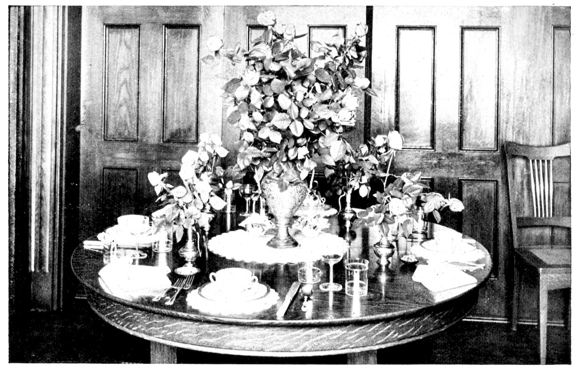 Table laid for Formal Luncheon. — A round oak table in a panelled room, with a tall vase of roses on a doily in the centre, smaller vases of roses around it, and places laid with cups on plates, napkins, glasses and silver.