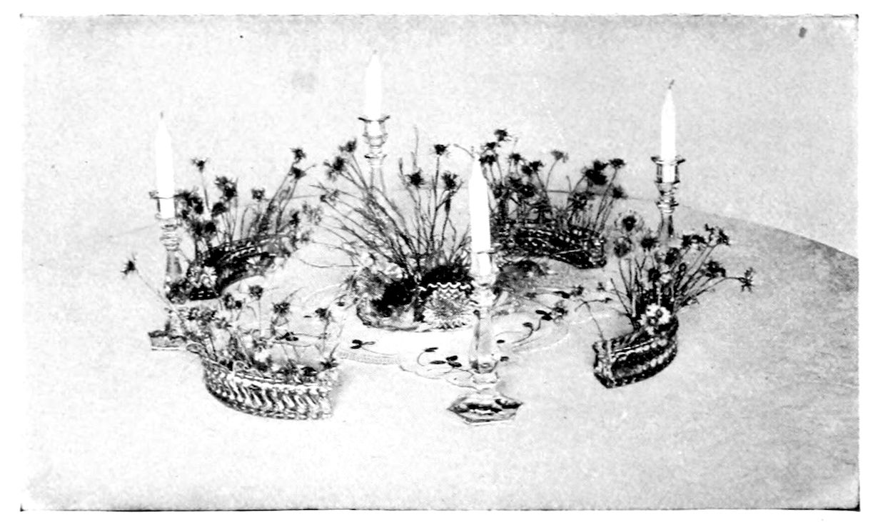 Centrepiece for Luncheon or Dinner Table. — Low oval baskets planted with slender-stemmed small flowers stand in a ring around a central bowl on a lace centrepiece, with four tall candles in glass candlesticks among them.
