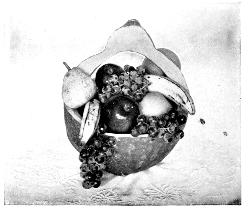 Centrepiece for Thanksgiving Dinner Table. — A hollowed shell cut into a basket with a tall handle, filled with pears, bananas, red apples, an orange and clusters of grapes, with one bunch hanging over the edge.