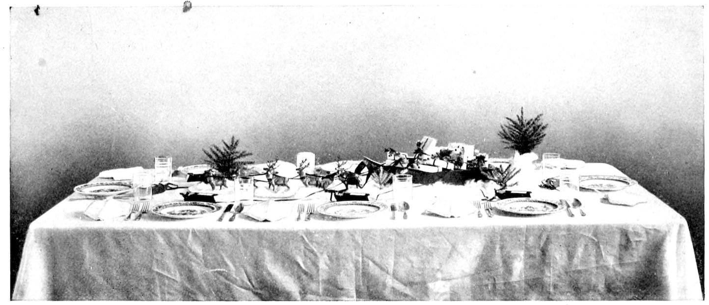 Christmas Dinner Table. — A long table laid with plates, glasses and folded napkins, with a toy sleigh piled with parcels drawn by a team of reindeer across cotton snow down the centre, and a small fir tree at each end.