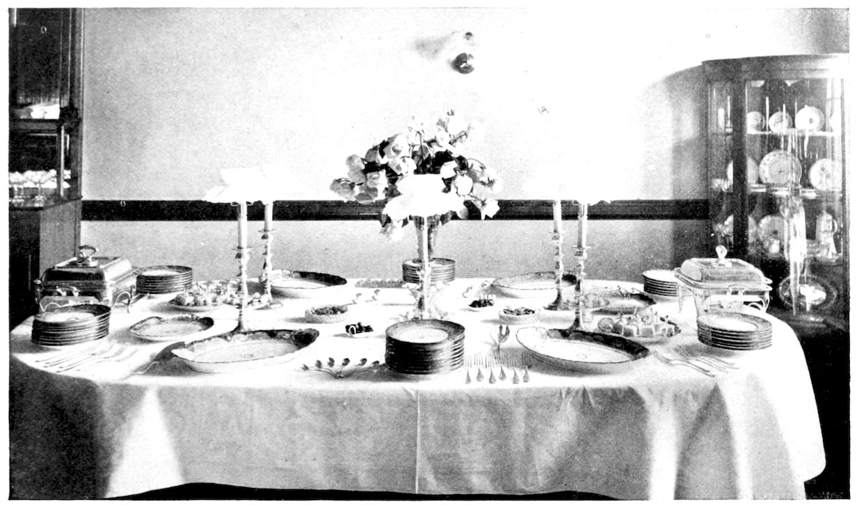 Table laid for Reception. — An oval reception table with a tall vase of roses in the centre, shaded candlesticks, chafing dishes at the ends, stacks of plates, platters, small dishes of sweets and rows of forks and spoons.