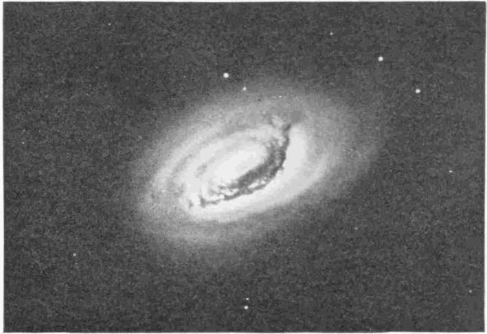 Another Spiral Nebula. Photo: G. W. Ritchey — An oval of soft light seen at a slant, with a bright centre crossed along one side by a dark, ragged lane of obscuring matter.