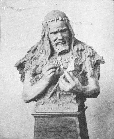 The Rutot Bust of a Cro-Magnon Man — A sculpted bust of a long-haired, bearded man wearing a skin cloak, a band of teeth in his hair and a necklace, working a small object with a pointed tool; the base reads “Homme Cro-Magnon”.