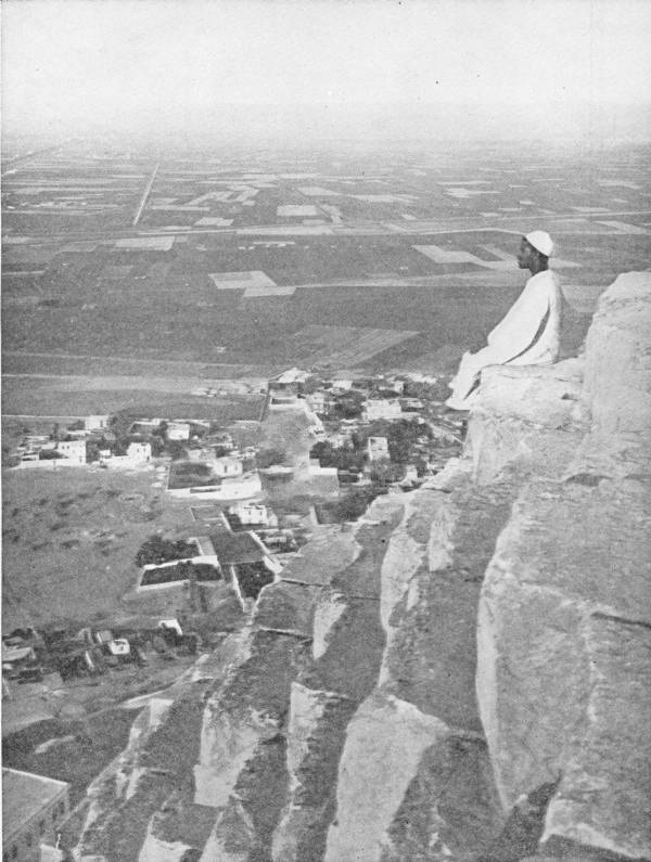 View from the Summit of the Great Pyramid of Cheops. Showing how these great monuments dominate the plain. Photo: D. McLeish — A man in white sits on a ledge of the pyramid’s great stone blocks, looking out over a village and a flat plain of fields and straight tracks stretching to a hazy horizon.