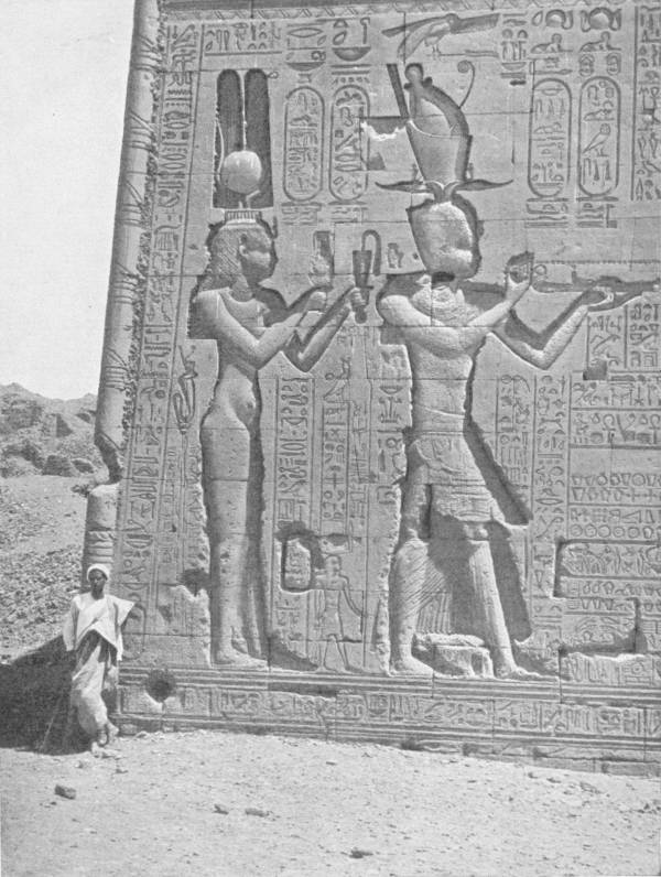 The Temple of Hathor at Dendereh. Photo: D. McLeish — A corner of the temple wall carved in relief with two tall figures, a crowned goddess and a crowned king holding out offerings, amid columns of hieroglyphs; a man in a turban stands at the foot of the wall.