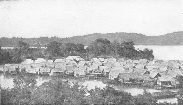 A Contemporary Lake Village. These Borneo dwellings are practically counterparts of the homes of European neolithic communities 6000 B.C. — A crowded village of thatched huts built on posts over the water of a bay, with trees in front and a line of wooded hills behind.