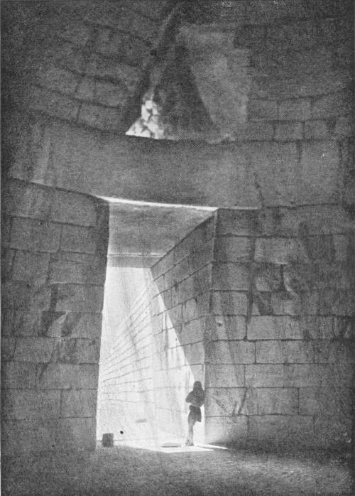 The Treasure House at Mycenæ. Photo: Fred Boissonnas — The inside of a round domed chamber of fitted stone blocks, looking toward its tall doorway, through which sunlight pours past a small figure standing in the passage beyond; a triangular opening shows above the lintel.
