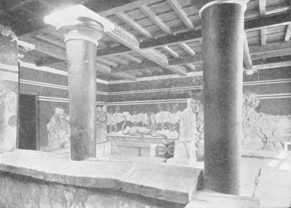 The Palace at Cnossos. The painted walls of the Throne Room. Photo: Fred Boissonnas — A room of dark tapering columns under a beamed ceiling, with painted walls behind showing a creature lying among tall plants beside a stone seat.