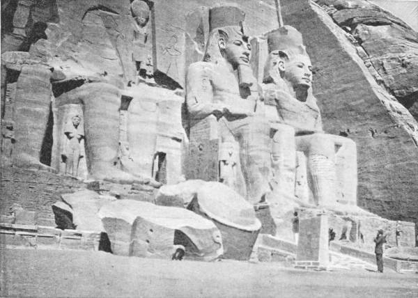 Temple at Abu Simbel. Showing the statues of Rameses II at entrance — Colossal seated statues carved out of the cliff face, wearing tall crowns, with smaller standing figures between their legs, fallen fragments in front and a man standing at the right for scale.