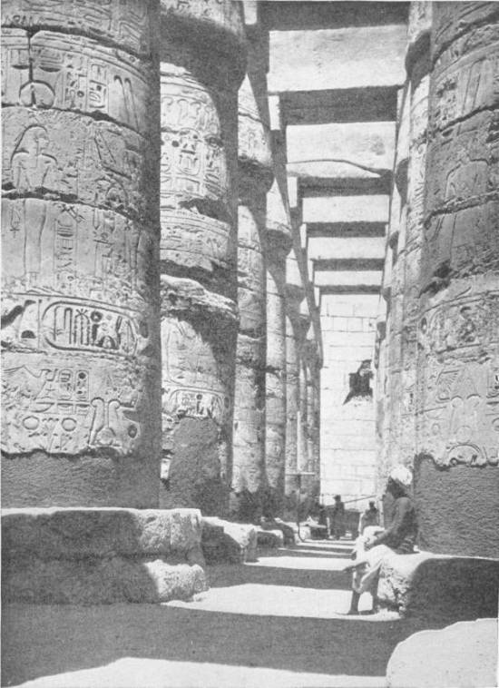 The Great Hypostyle Hall at Karnak. Photo: D. McLeish — A narrow aisle between enormous columns carved all over with figures and hieroglyphs, roofed by stone beams, with a man seated at the foot of one pillar.