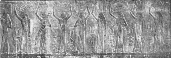 Frieze Showing Egyptian Female Slaves Carrying Luxurious Foods. Photo: Jacques Boyer — A carved relief of a row of women walking in single file, each carrying a basket or tray on her head, with hieroglyphic signs between them.
