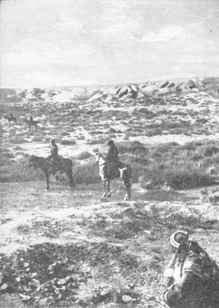 The Mound of Nippur. The site of a city which recent excavations have proved to date from at least as early as 5000 B.C., and probably 1000 years earlier. Photo: Underwood & Underwood — A low, bare landscape of scrub and earth mounds, with horsemen riding across it and a figure in a headcloth crouching in the foreground.