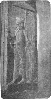 Persian Monarch. From the ruins of Persepolis. Photo: Miss F. Biggs — A stone relief of a bearded king in a long robe walking to the right, with a smaller attendant behind him holding up the staff of a parasol.