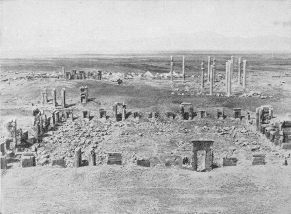 The Ruins of Persepolis. The capital city of the Persian Empire; burnt by Alexander the Great. Photo: Major W. F. P. Rodd — A wide stony plain strewn with the ruins of Persepolis: rows of broken doorways and wall footings in the foreground, and a cluster of tall lone columns beyond, with hills on the horizon.