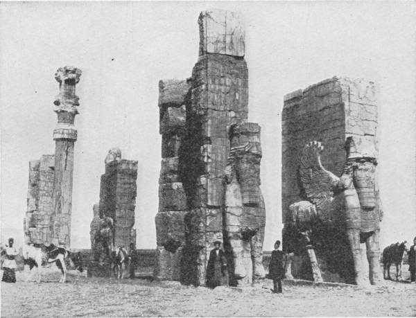 The Great Porch of Xerxes, at Persepolis. Photo: Major W. F. P. Rodd — Massive ruined stone gate-towers, their sides carved with huge winged bulls, beside a single tall column; men in long coats and a few horses stand at their feet for scale.