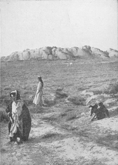 The Mound at Babylon. Beneath which are the remains of a great palace of Nebuchadnezzar. Photo: Underwood & Underwood — A long, low, eroded mound rising from a flat grassy plain, with three figures in robes and a seated man in a straw hat in the foreground.