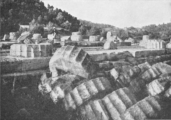 Ruins of the Great Temple of Zeus at Olympia. Photo: Fred Boissonnas — Fallen column drums of the temple lying in a tumbled row across its stone platform, their fluting still sharp, with wooded hills behind.