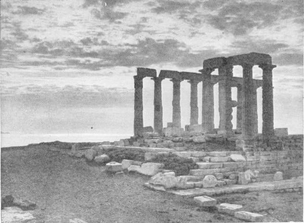 The Temple of Neptune (Poseidon) at Cape Sunium. Photo: Fred Boissonnas — A ruined temple of slender columns standing on a headland against a cloudy sky, with the sea beyond and fallen blocks in the foreground.