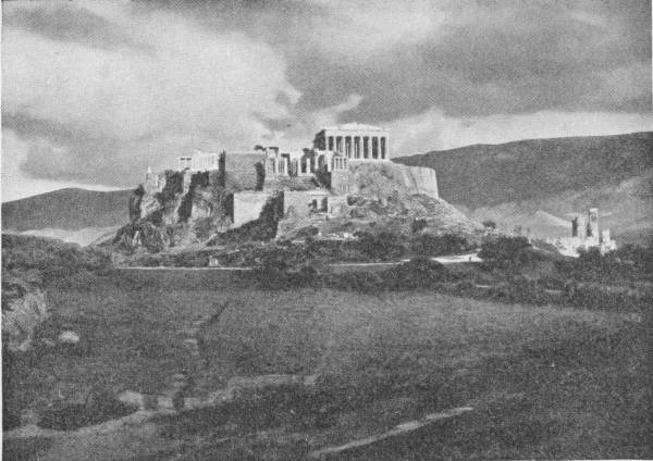 The Acropolis, Athens. The marvellous group of Temples and monuments built under the inspriration of Pericles. Photo: Fred Boissonnas — The rocky hill of the Acropolis seen across open fields, crowned by its walls and the columns of the Parthenon, under a heavy clouded sky.