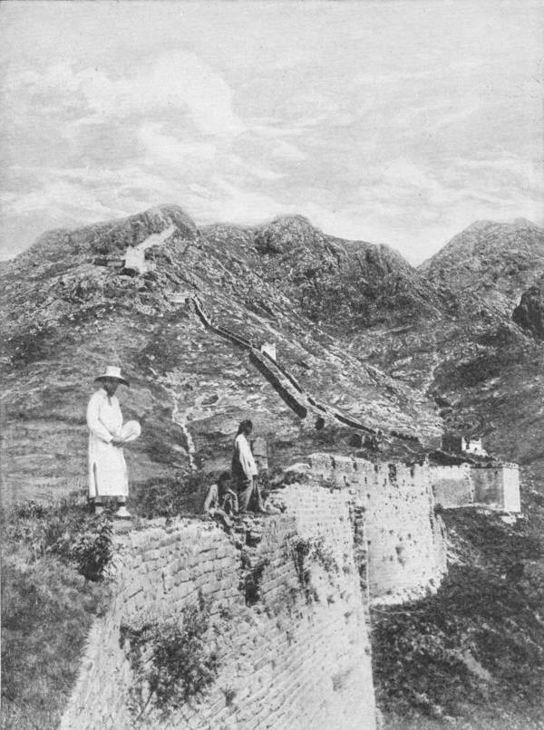 The Great Wall of China. As it crosses the mountains in Manchuria. Photo: Underwood & Underwood — The Great Wall climbing steeply over bare mountain ridges in a line of battlements and watch-towers, with a man in white and two others standing on the near section.
